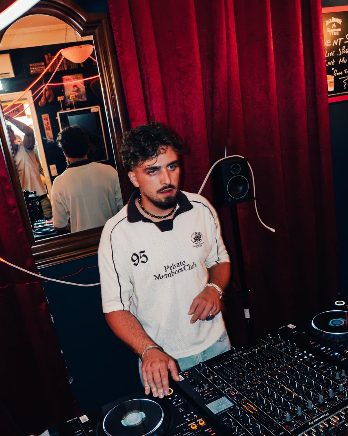 A DJ in a white football shirt at the decks, red curtains behind him