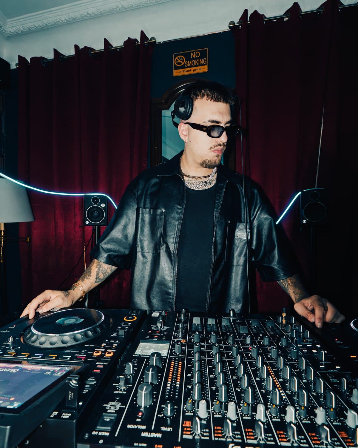 A DJ in a leather shirt and sunglasses at the decks, red curtains behind him