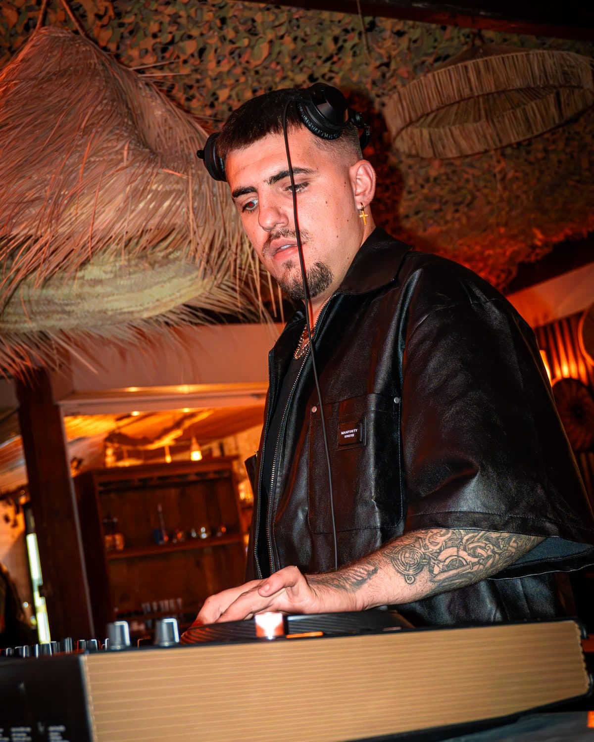 A DJ in a leather shirt at the decks under the straw roof of Le Vieux Rocher, warm light
