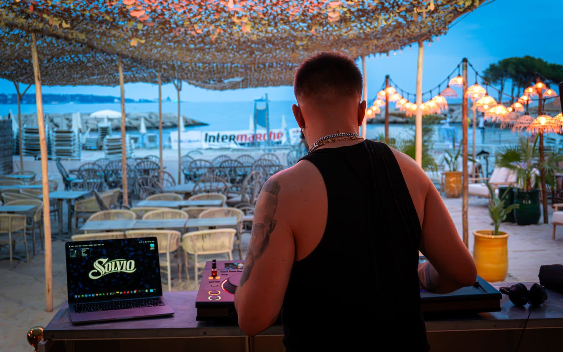 Solvio at the decks at Le Vieux Rocher, seen from behind, his logo on the laptop, the beach and the sea in the background